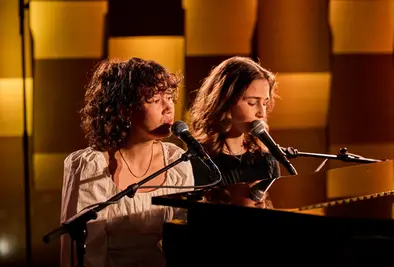 Zwei junge Frauen singen am Mikrofon, eine von ihnen spielt Klavier.