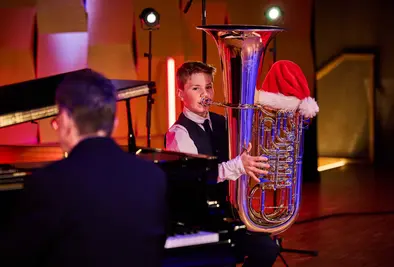 Ein Junge spielt Tuba auf einer Bühne, auf dem Instrument steckt eine Weihnachtsmütze. Ein Pianist begleitet ihn.