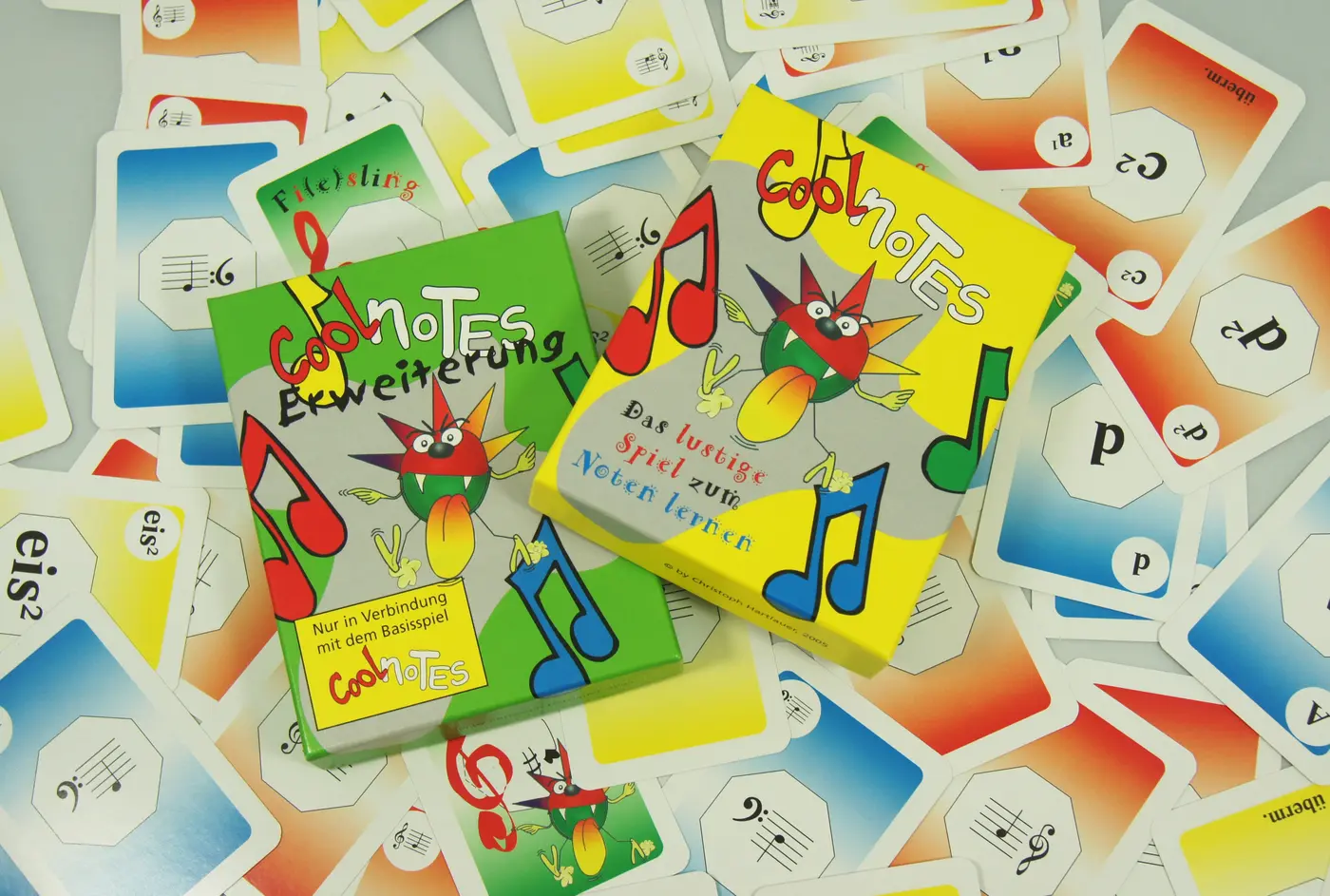 Das gelbe Basisspiel „Cool Notes“ und die grüne Erweiterung liegen auf einer Fläche voller einzelner Spielkarten.
