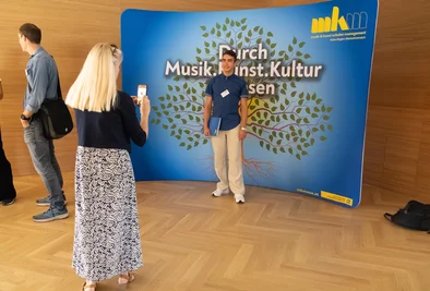Teilnehmer wird vor einer Veranstaltungswand mit dem Schriftzug „Durch Musik. Kunst. Kultur“ fotografiert.
