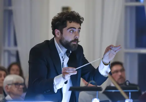 Ein Mann dirigiert ein Orchester, im Hintergrund ist das Publikum zu sehen.