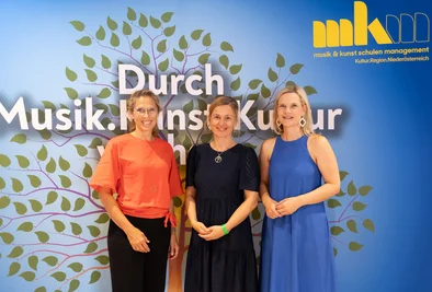 Drei Personen stehen gemeinsam vor einer Veranstaltungswand mit dem Schriftzug „Durch Musik. Kunst. Kultur“.