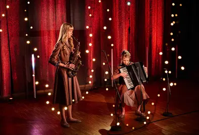 Zwei Mädchen spielen auf einer Bühne, eines am Saxofon und eines am Akkordeon, umgeben von warmen Lichtpunkten.