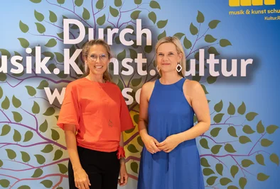 Zwei Personen stehen gemeinsam vor einer Veranstaltungswand mit dem Schriftzug „Durch Musik. Kunst. Kultur“.