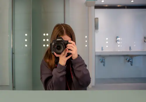 Ein Mädchen hält eine Kamera vor ihr Gesicht und macht ein Spiegelselfie in einer öffentlichen Toilette.
