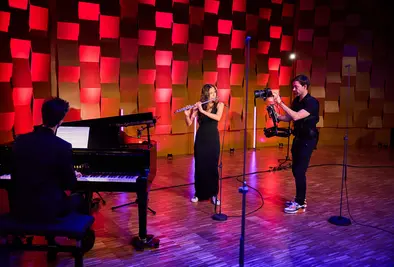 Eine Flötistin spielt auf einer Bühne, begleitet von einem Pianisten. Ein Kameramann filmt die Szene mit einer stabilisierten Kamera. Der Hintergrund ist in rotes Bühnenlicht getaucht und zeigt strukturierte Wandpaneele.