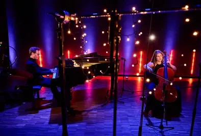 Eine Cellistin und ein Pianist spielen auf einer stimmungsvoll beleuchteten Bühne. Zwischen den beiden hängen Lichterketten und Mikrofone, die Szene erscheint warm und festlich ausgeleuchtet.