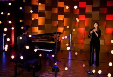 Eine Oboistin spielt auf einer Bühne, begleitet von einem Pianisten. Vor der Szene hängen unscharfe Lichter.