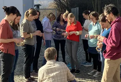 Teilnehmende stehen im Freien im Halbkreis um eine hockende Person.