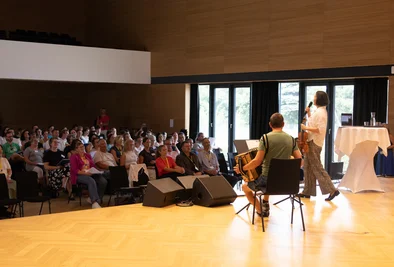 Musikerinnen und Musiker stehen auf der Bühne vor dem sitzenden Publikum.