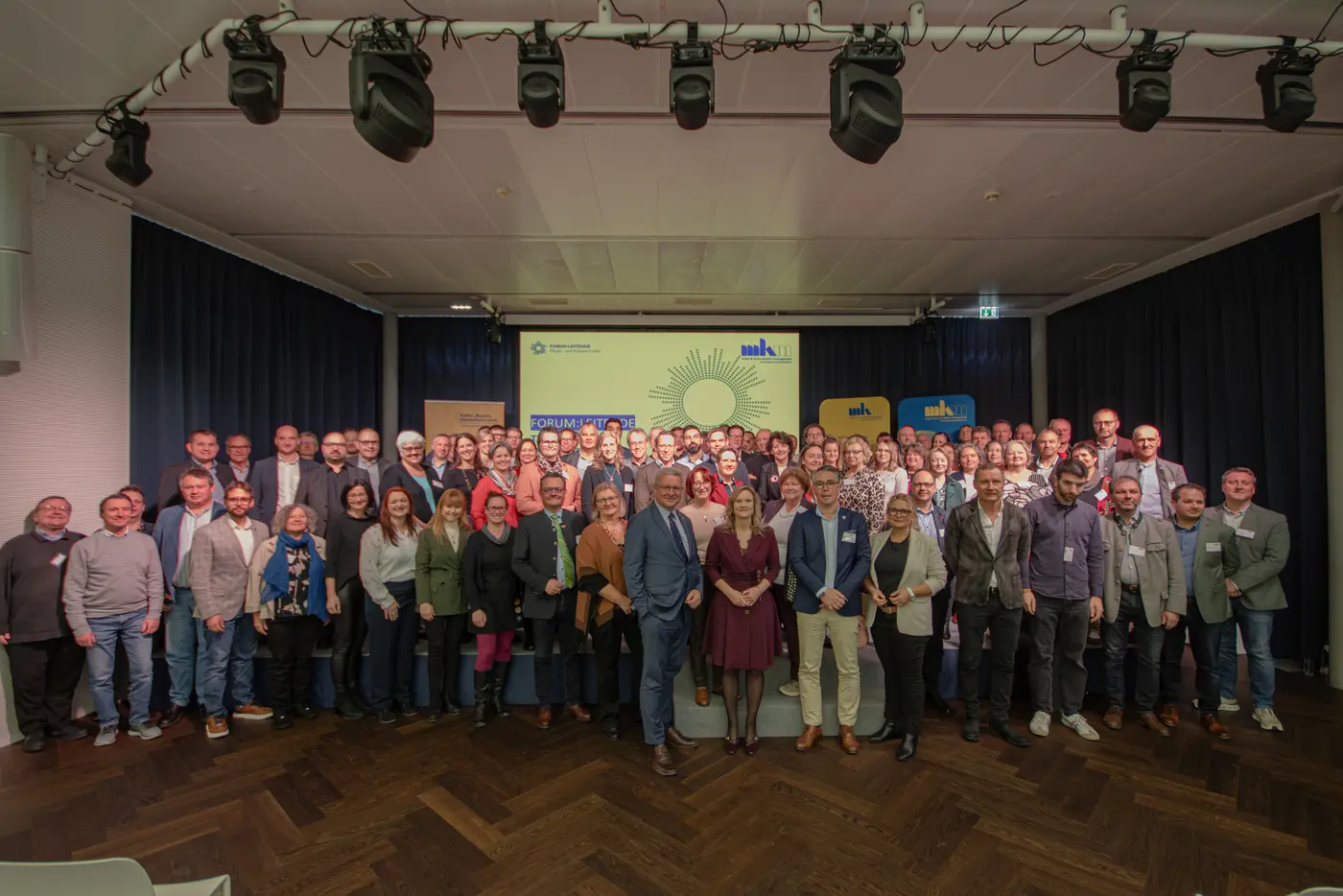 Gruppenfoto aller Teilnehmer eines Arbeitstreffens auf einer Bühne, im Hintergrund eine Präsentation auf einer Leinwand.