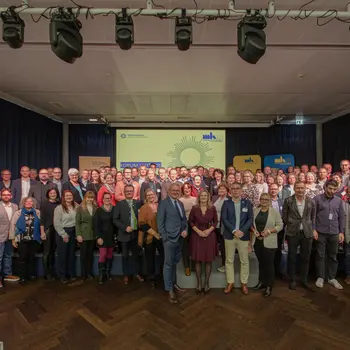 Gruppenfoto aller Teilnehmer eines Arbeitstreffens auf einer Bühne, im Hintergrund eine Präsentation auf einer Leinwand.