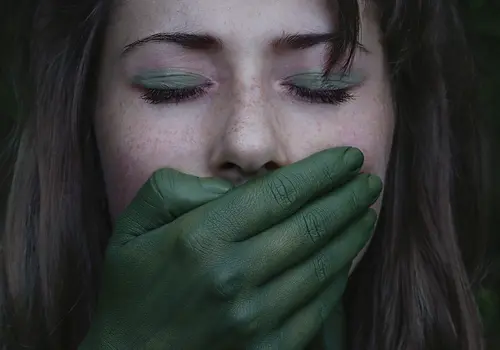 Eine Frau mit geschlossenen Augen hält sich mit einer grün bemalten Hand den Mund zu, während die andere ihren Hals umfasst. Auf ihrem Kopf trägt sie einen Kranz aus grünen Blättern.