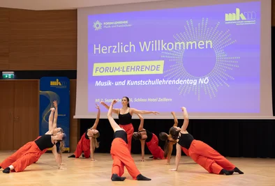 Tanzgruppe bei einer Bühnenperformance vor einer Präsentationsfolie mit der Begrüßung zum Lehrendentag.