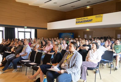 Publikum sitzt im Veranstaltungssaal und verfolgt eine Präsentation.