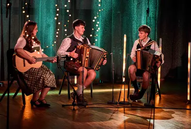 Drei junge Menschen in Tracht spielen auf der Bühne Gitarre und Steirische Harmonikas.