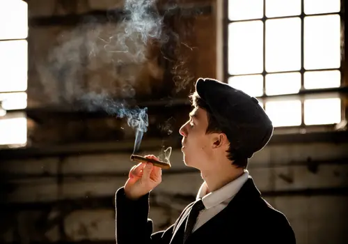 Eine Person in vintage-inspirierter Kleidung mit Mütze steht in einem alten Industriegebäude und raucht eine Zigarre. Rauch steigt vor einem großen, hellen Fenster nach oben.