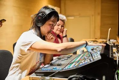 Eine Frau mit Kopfhörern bedient konzentriert ein Mischpult, während eine zweite Person daneben zusieht.