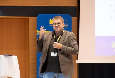 Mann mit Mikrofon spricht auf der Bühne neben einem Präsentationsbildschirm.
