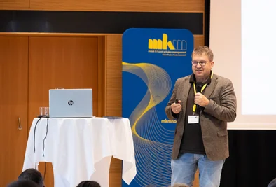Redner mit Mikrofon steht neben einem Laptop und einem Präsentationsbildschirm.