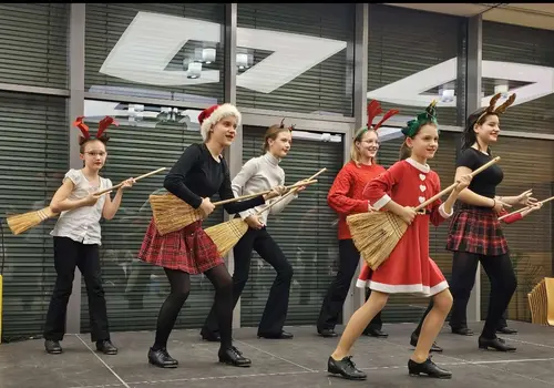 Kinder mit weihnachtlichem Kopfschmuck führen etwas mit Besen in den Händen auf.