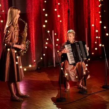 Links steht ein Mädchen, das Saxophon spielt. Rechts daneben sitzt ein weiteres Mädchen und spielt Akkordeon. Im Hintergrund hängt ein roter Vorhang, im Vordergrund sind weiß leuchtende Punkte zu sehen.