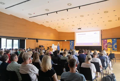 Teilnehmerinnen und Teilnehmer verfolgen eine Präsentation im hellen Veranstaltungssaal.