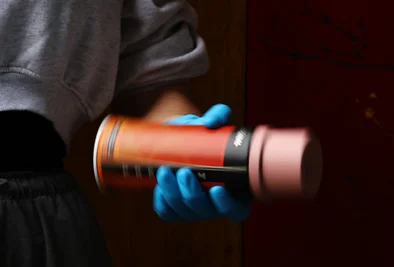eine Hand mit blauen Handschuhen hält eine Spraydose vor dunklem Hintergrund