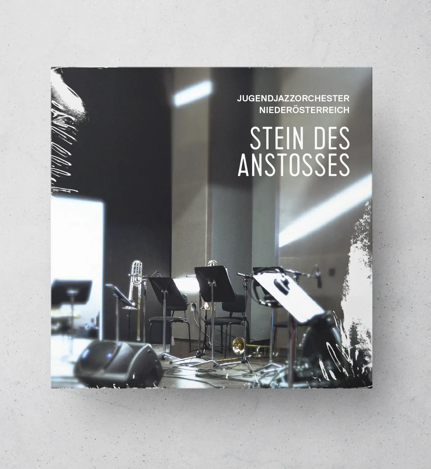 CD-Hülle mit einem Foto eines leeren Proberaums mit Instrumenten und Notenständern. Oben rechts steht der Text: „Jugendjazzorchester Niederösterreich – Stein des Anstoßes“.