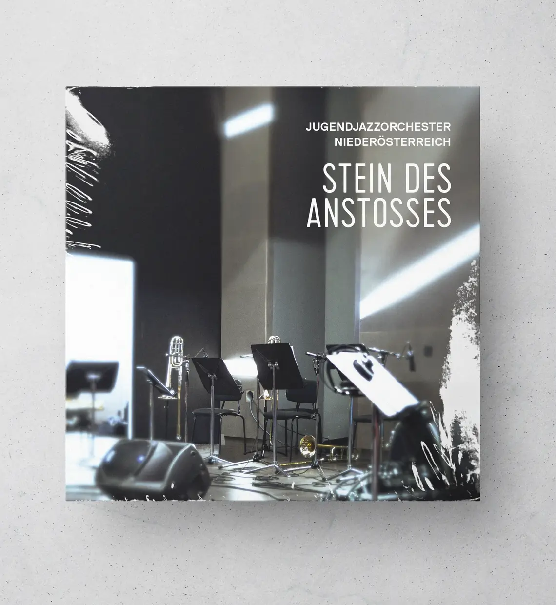 CD-Hülle mit einem Foto eines leeren Proberaums mit Instrumenten und Notenständern. Oben rechts steht der Text: „Jugendjazzorchester Niederösterreich – Stein des Anstoßes“.