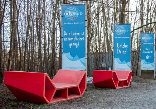 Drei große rote Sitzskulpturen stehen im Freien vor einer Reihe blauer Werbebanner der „odyssee“ Bar, Bistro und Bootsverleih. Die Banner zeigen Slogans wie „Dein Leben ist unkompliziert genug!“, „Erlebe Deine!“ und „Hart Steuerbord!“. Im Hintergrund stehen kahle Bäume.