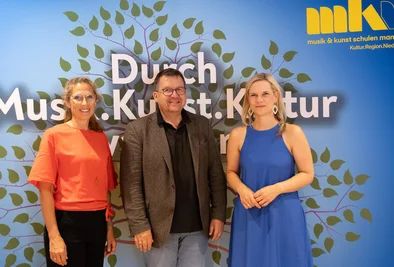 Drei Personen stehen gemeinsam vor einer Veranstaltungswand mit dem Schriftzug „Durch Musik. Kunst. Kultur“.