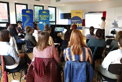 Teilnehmende sitzen im Seminarraum vor einer Vortragenden; rechts ein KUKUDU-Banner.