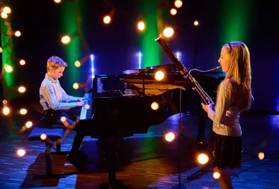 Ein Junge spielt Klavier, während ein Mädchen Fagott spielt. Vor der Bühne hängen unscharfe Lichter.