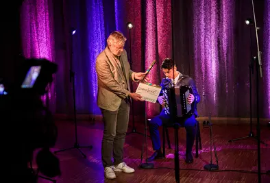 Ein Mann hält eine Filmklappe, während ein junger Musiker am Akkordeon auf der Bühne sitzt.