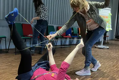 Eine Person liegt auf dem Rücken am Boden während eine Andere Wolle um sie wickelt.