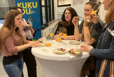 Fünf Frauen stehen an einem Stehtisch und trinken Sekt und essen Brötchen.