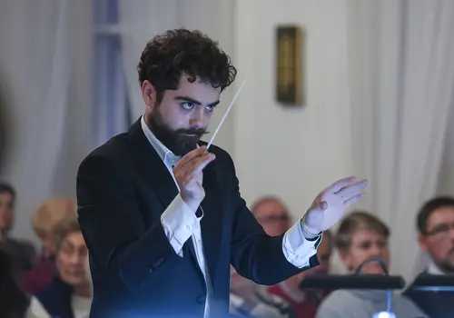 Ein Mann dirigiert ein Orchester, im Hintergrund ist das Publikum zu sehen.