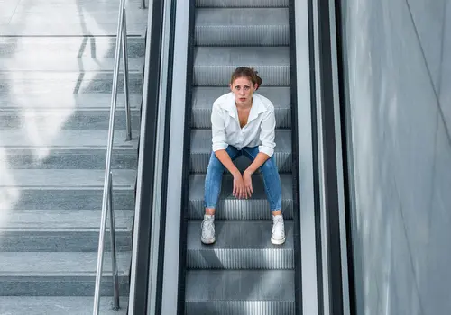 Eine Person sitzt mittig auf einer Rolltreppe während sie ernst in die Kamera schaut.