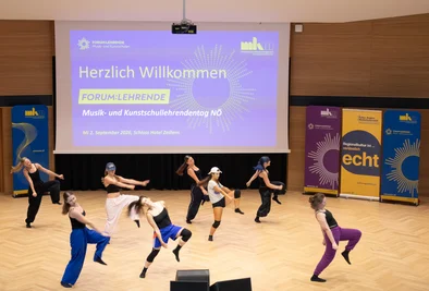 Tanzende Personen in verschiedenen Farben auf der Bühne vor einer Präsentationsfolie.